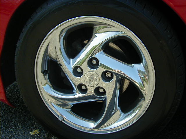 Chrome swirl-spoke alloy wheel with Ford center cap on a red car