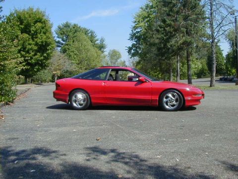 1997 Ford Probe - A red 1997 Ford Probe is parked on a paved surface with trees in the background under a clear blue sky.