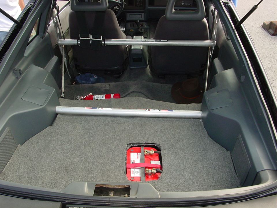 Car interior showing front seats, roll bar, fire extinguisher, battery in trunk, and a hat
