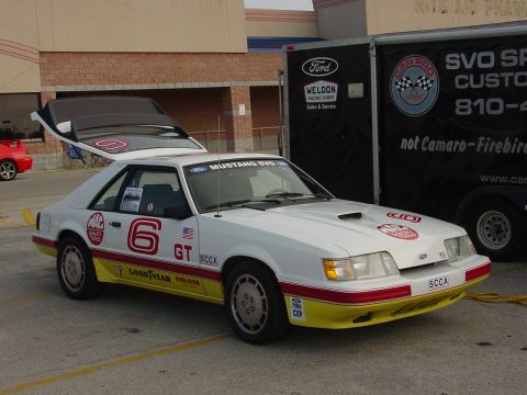 1986 Ford Mustang - A white Ford Mustang GT race car with racing decals is parked next to a trailer at an event.