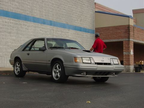 1986 Ford Mustang - A silver coupe with a hood scoop is parked in front of a brick building, with a person leaning on the hood.