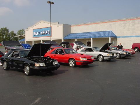 1986 Ford Mustang - A line of Ford Mustangs, including a black coupe with its hood raised, are parked outside a building.