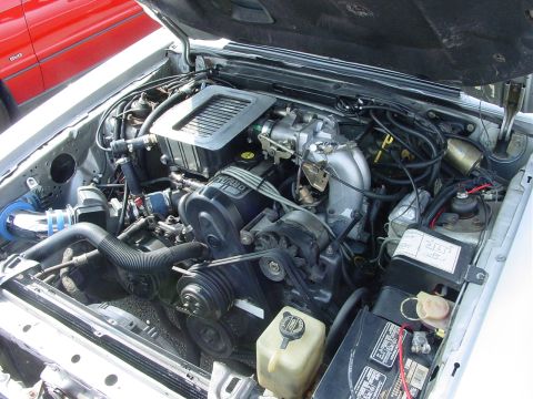 1986 Ford Mustang - The engine bay of a 1986 Ford Mustang is visible, with a red car in the background.
