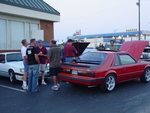 1986 Ford Mustang - A red Ford Mustang is parked with its hood open at a car meet, with other vehicles and people around.