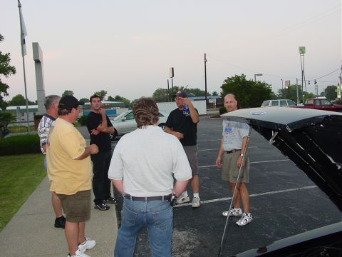 1986 Ford Mustang - A group of men are gathered around a black car with its hood open in a parking lot.
