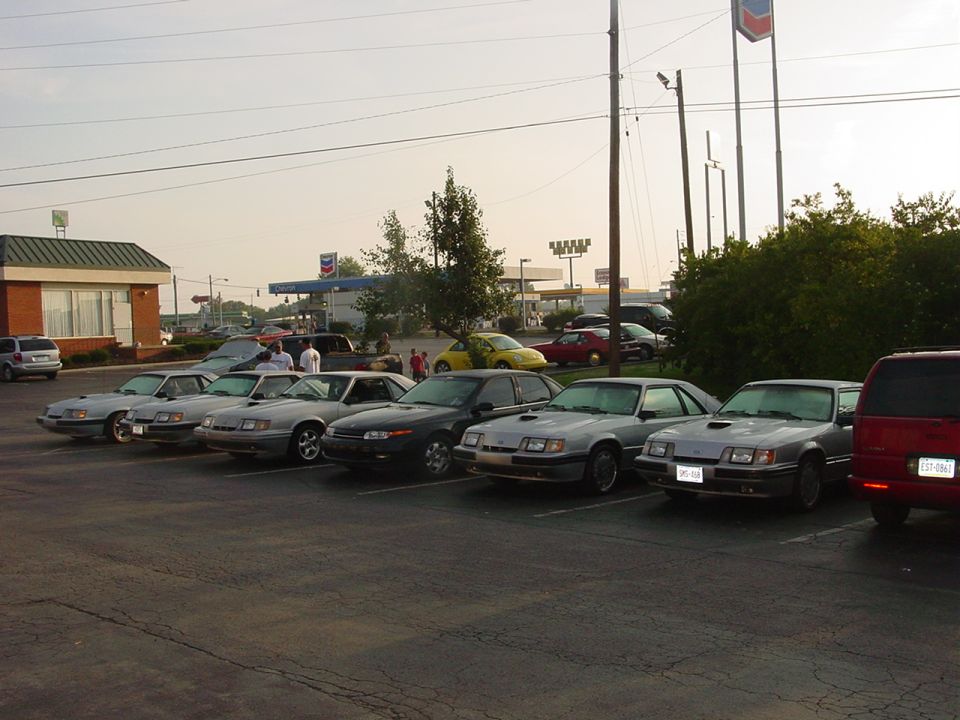 Several 1987 Ford Mustangs parked in a lot with a gas station in the background