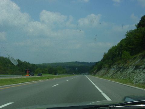 1986 Ford Mustang - A view from a vehicle driving on a highway surrounded by green hills and trees under a cloudy sky.
