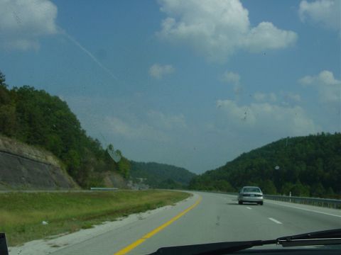 1986 Ford Mustang - A silver sedan travels down a highway, surrounded by lush green hills under a cloudy sky.