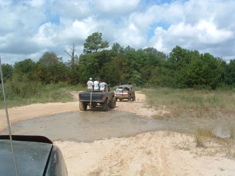 1993 Ford Bronco - Several people ride in the back of trucks as they navigate a muddy trail through a wooded area.