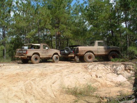 1993 Ford Bronco - Two muddy Ford trucks, a Bronco and a pickup, are parked on a dirt path surrounded by trees.