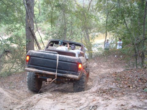 1993 Ford Bronco - Off-road vehicle with passengers drives through a muddy trail surrounded by trees.