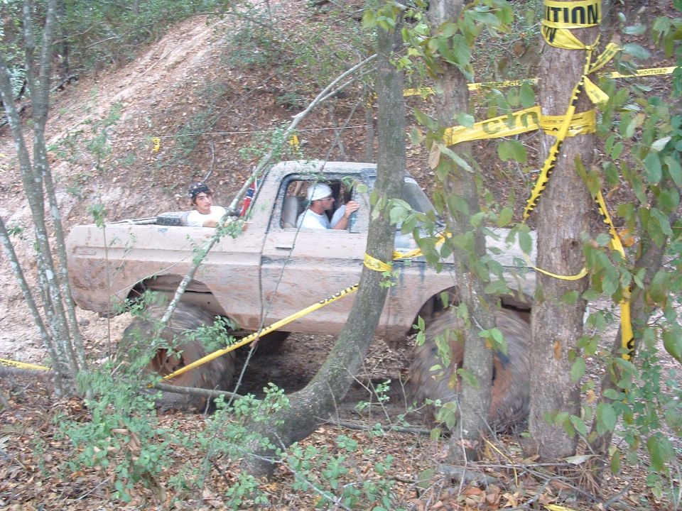 Muddy 1980s pickup truck with large tires, side view, with people in the cab and bed