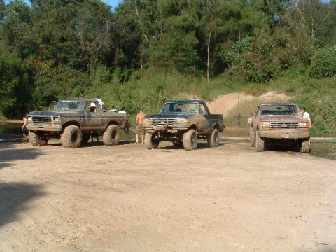 1993 Ford Bronco - Several lifted trucks are parked off-road near a body of water and wooded area.