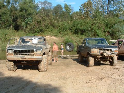 1993 Ford Bronco - Two lifted Ford trucks are parked near water, with people in the background. The black truck has a 'JEEP RESCUE TEAM' banner.