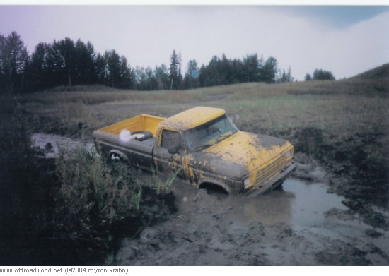 Yellow vintage Ford pickup stuck axle-deep in a muddy field bog