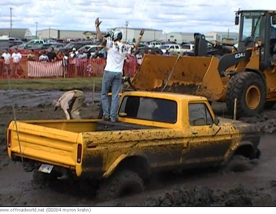 Person standing in bed of mud-covered yellow pickup as a wheel loader approaches