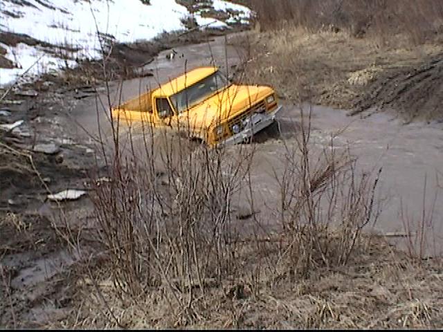 Yellow pickup truck driving through a muddy water-filled trail with snow on the banks