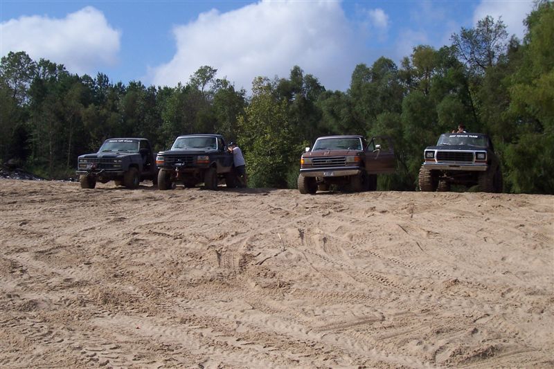 Four trucks parked on a sandy hill with trees in the background. One truck has a person standing next to it