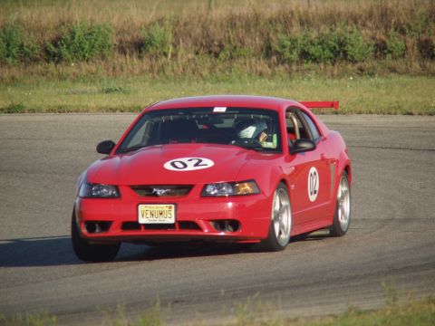 2000 Ford Mustang - A red 2000 Ford Mustang with racing decals and a roll cage is seen on a race track.