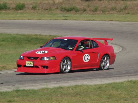 2000 Ford Mustang - This red 2000 Ford Mustang, identified by its body style and racing decals, is captured on a race track.