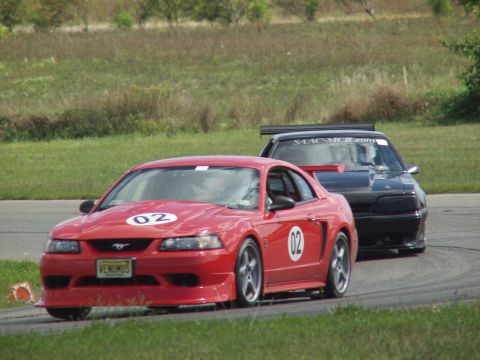2000 Ford Mustang - Two cars race on a track, with a red Mustang in the foreground and a black car behind it.