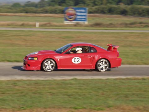 2000 Ford Mustang - A red 2000 Ford Mustang coupe, identified by its body style and racing number, is captured in a side profile on a race track.