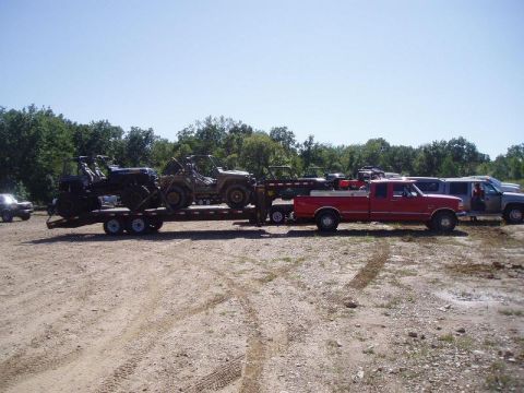 1992 Ford Bronco - A red pickup truck tows a trailer loaded with ATVs and a Jeep-like vehicle, with other vehicles parked nearby.