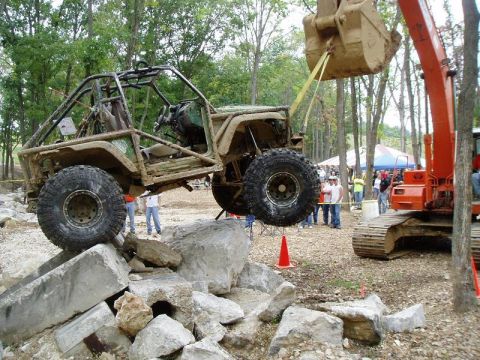 1992 Ford Bronco - An excavator lifts a heavily modified off-road vehicle over a rocky obstacle course.