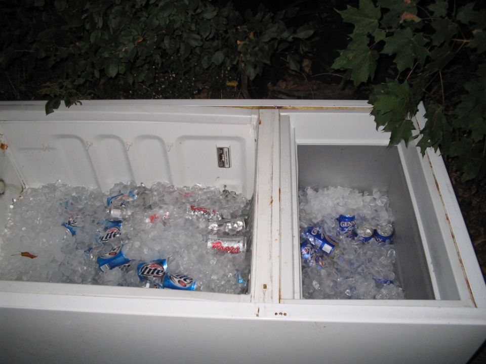 White cooler packed with ice and beer cans, sitting outside near some trees