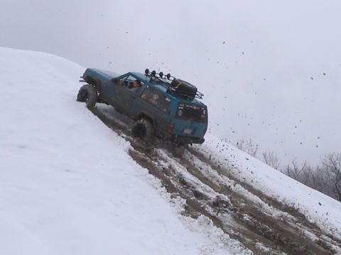 1992 Ford Bronco - A blue SUV navigates a challenging snowy and muddy slope, kicking up dirt and snow.