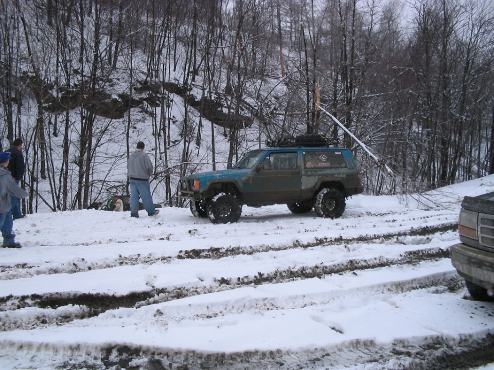 Modified Jeep Cherokee with large tires parked in snow, another vehicle partially visible on the right