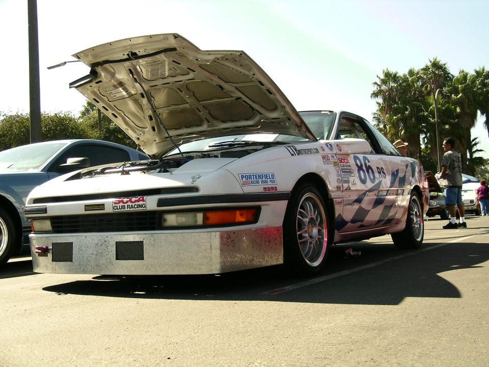 White race car with hood up, number 86 on door, checkered flag pattern on side. Another car parked next to it
