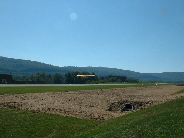 Yellow airplane in flight over a field, with a building and mountains behind it