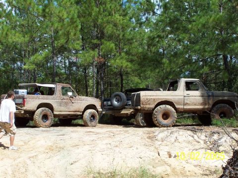 1987 Ford Bronco - Two Broncos, one with a removable top, are covered in mud on a dirt trail.