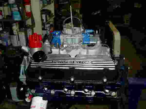 Ford V8 engine with black valve covers reading "POWERED BY FORD", red distributor cap, and blue ignition components