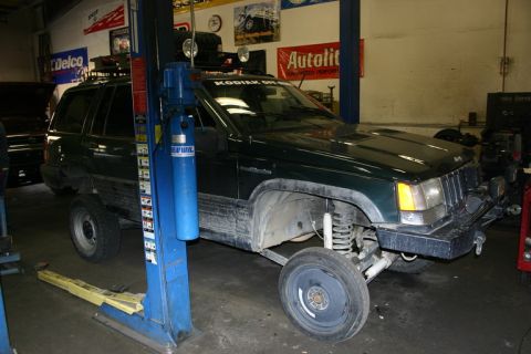 2003 Jeep TJ - A dark green Jeep Grand Cherokee is shown on a lift in a garage, with its front passenger wheel removed for service.