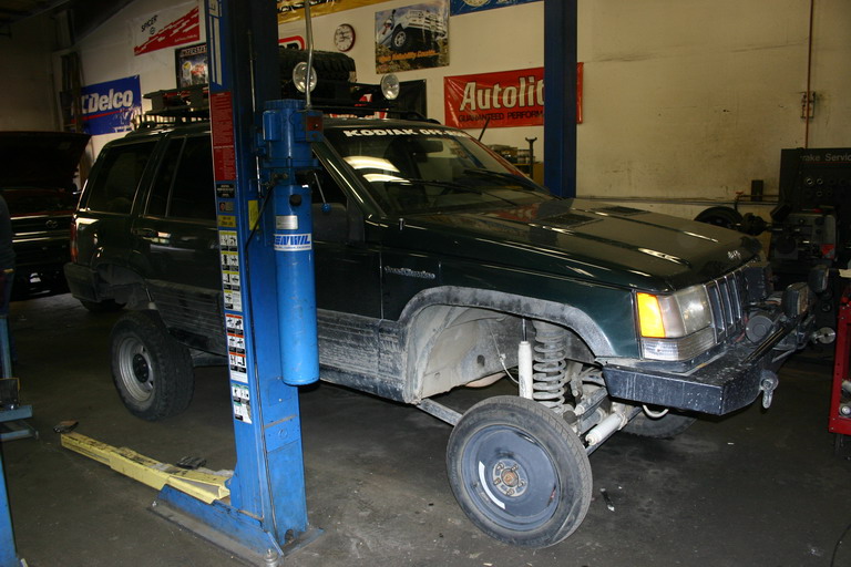 Dark green Jeep Grand Cherokee on a lift with one wheel removed. "KODIAK OFF-ROAD" is on the windshield
