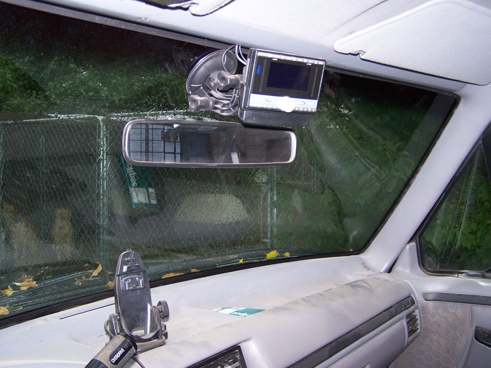 Vehicle interior showing dashboard, rearview mirror, and a mounted monitor. Some leaves are on the windshield