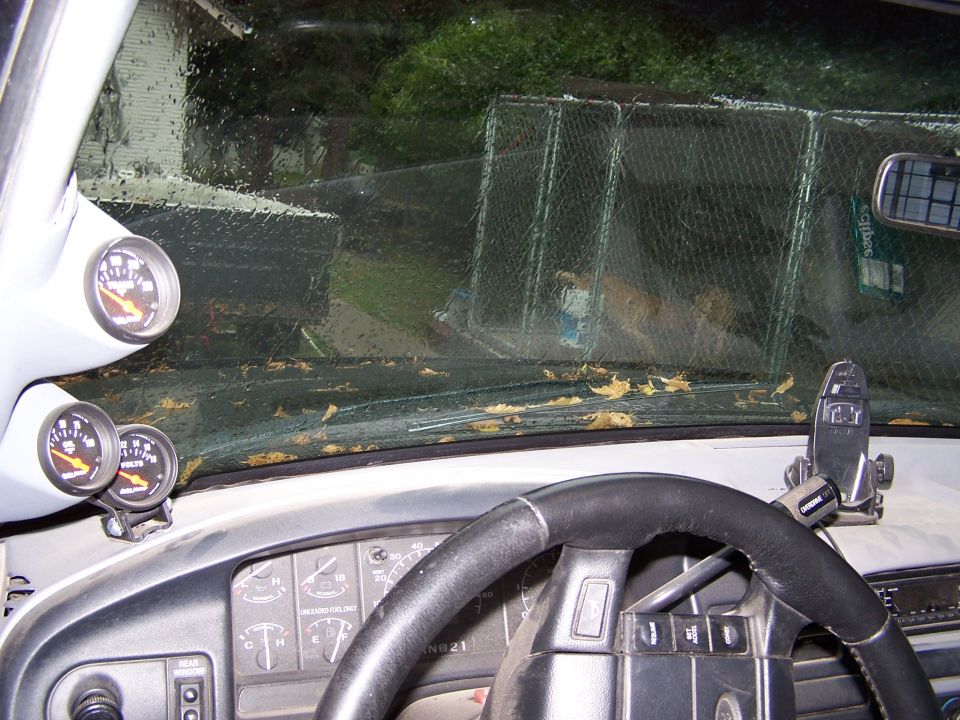 Interior shot of a vehicle's dashboard with two gauges mounted on the A-pillar and a steering wheel. The windshield is dirty with leaves