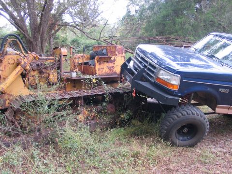 1995 Ford Bronco - A blue Ford Bronco is parked next to a large, weathered bulldozer in an overgrown outdoor area.