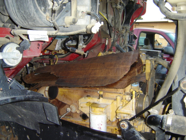 Engine compartment with yellow engine block, brown damaged insulation, and red cab. A red pickup truck is visible in the background