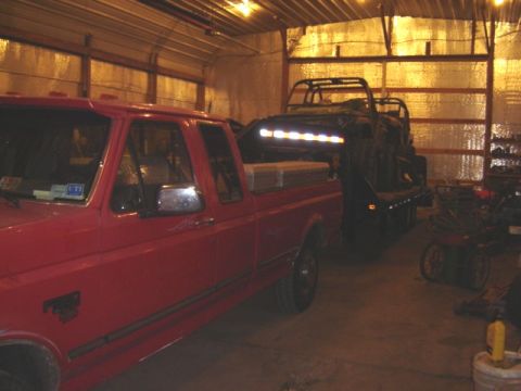 1992 Ford Bronco - A red pickup truck is parked inside a garage next to a trailer with a vehicle on it.