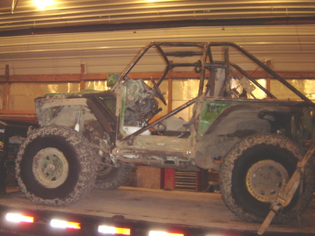 Mud-covered off-road buggy with custom roll cage, missing body panels, on a trailer