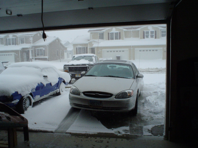 White Ford Taurus and snow-covered car beside pickup, viewed from inside garage during heavy snow