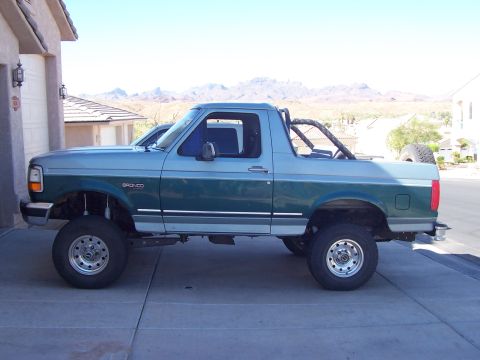 1996 Ford Bronco - Side profile of a two-tone green-over-silver full-size Ford Bronco with the removable hardtop off, exposing the interior roll bar, parked l…