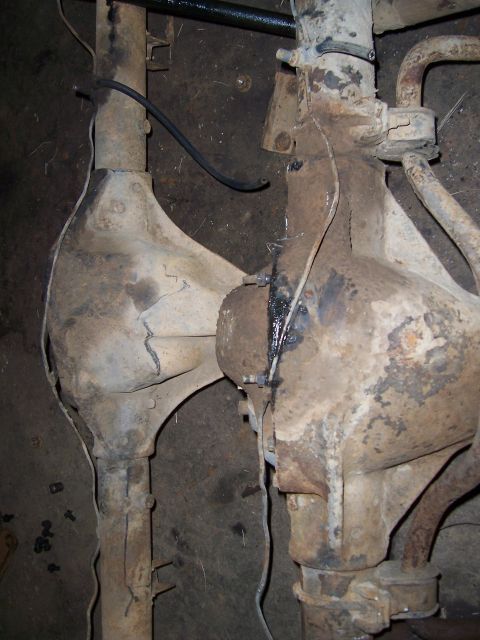 1983 Ford Bronco - Two rusty solid axle housings lying side by side on a dirt shop floor, both stripped down to bare cast center sections with dried grease ar…