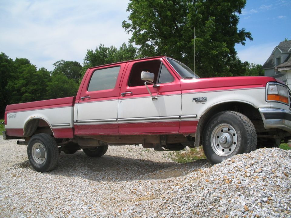 Red and white Ford F-350 crew cab pickup truck parked on gravel with a house and trees in the background