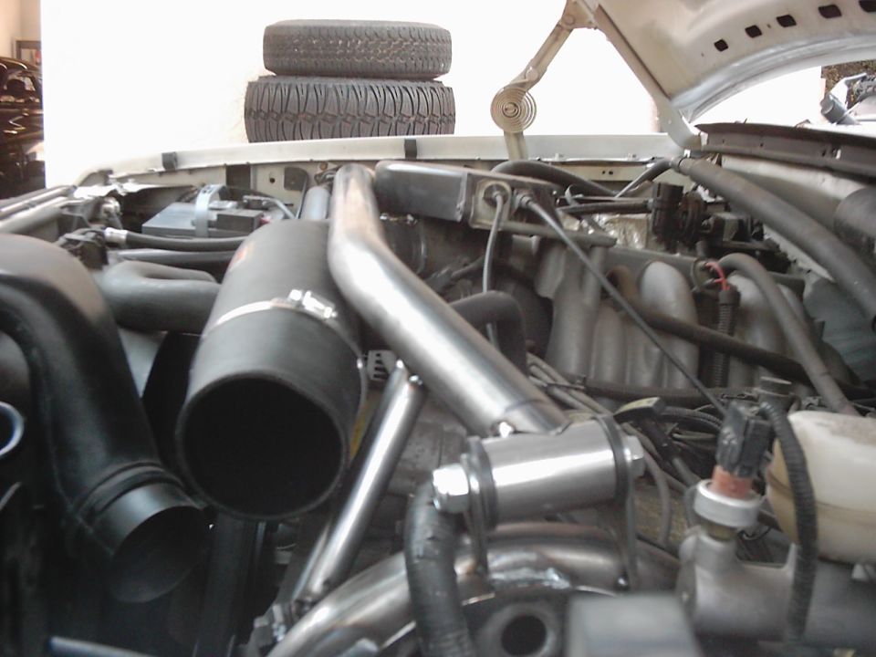Engine bay with large diameter intake pipes, custom tubing, and a battery. Two tires are stacked on top