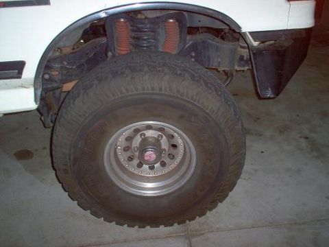 1988 Ford Bronco - Close-up of the front wheel assembly of a white truck, showing the suspension and tire.