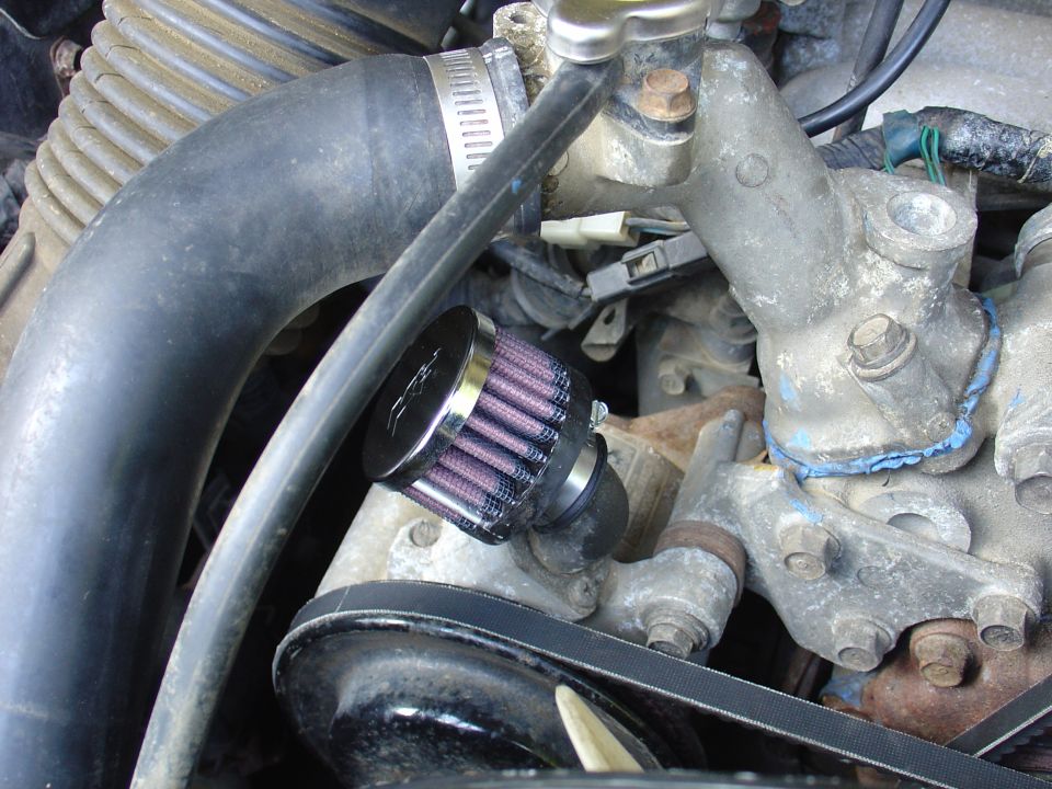 Close-up of engine bay showing a small purple air filter attached to a hose, with various metal parts and a black belt visible
